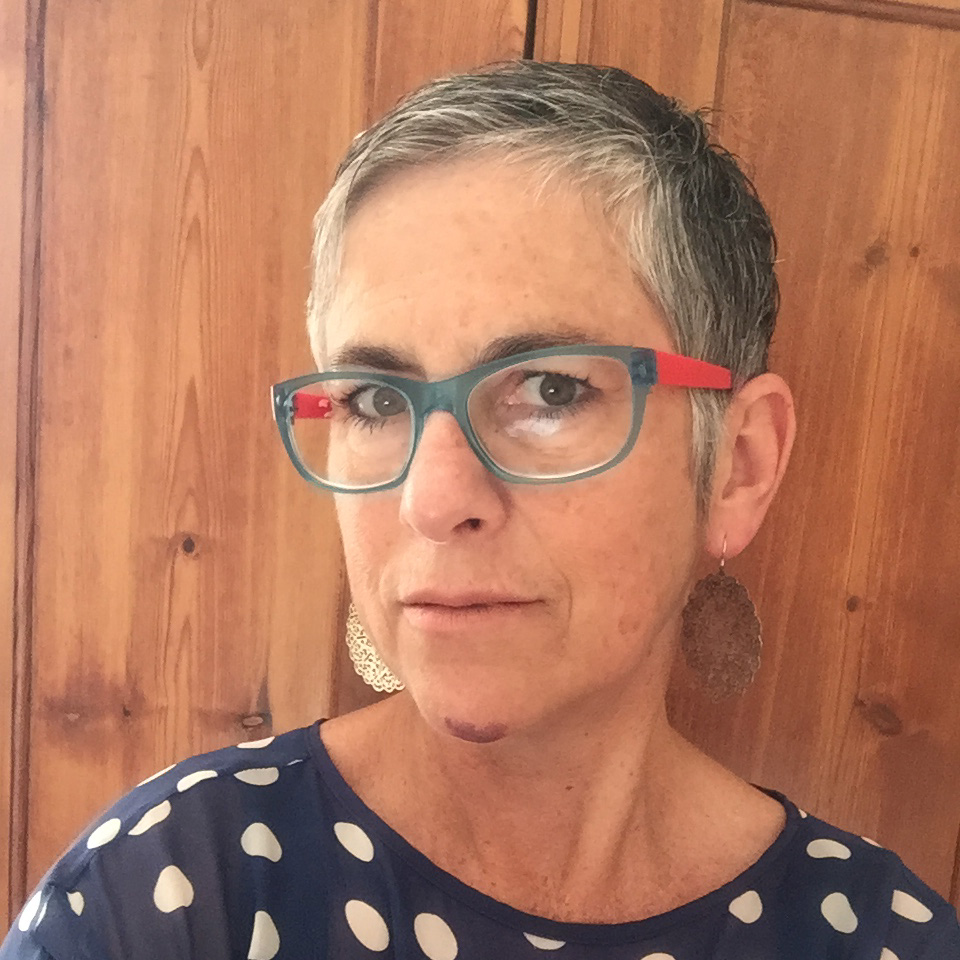 Woman with short grey hair, red and grey rimmed glasses and a blue and white polka dot shirt.