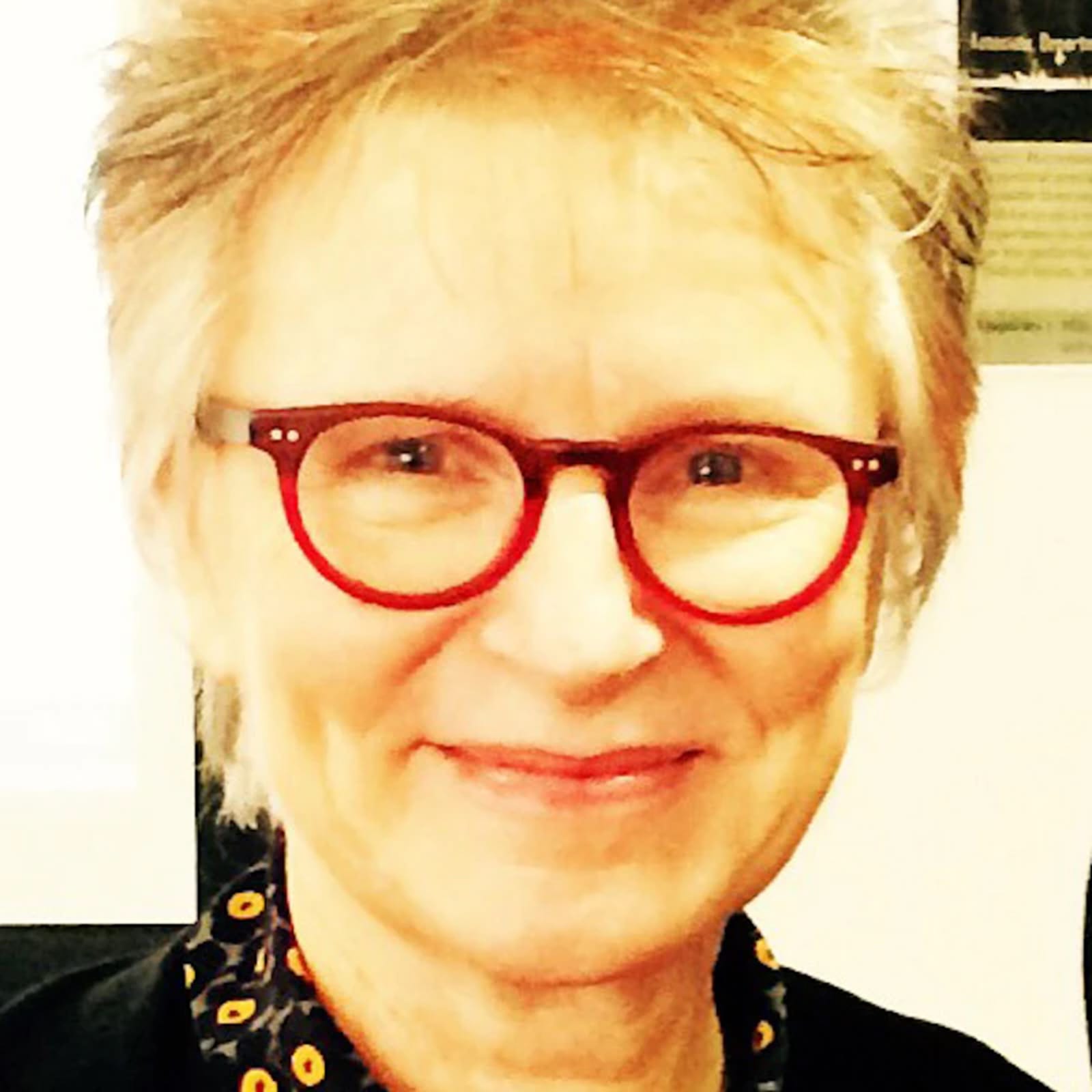 close-up of womans face with short blond-grey hair and red rimmed glasses, smiling